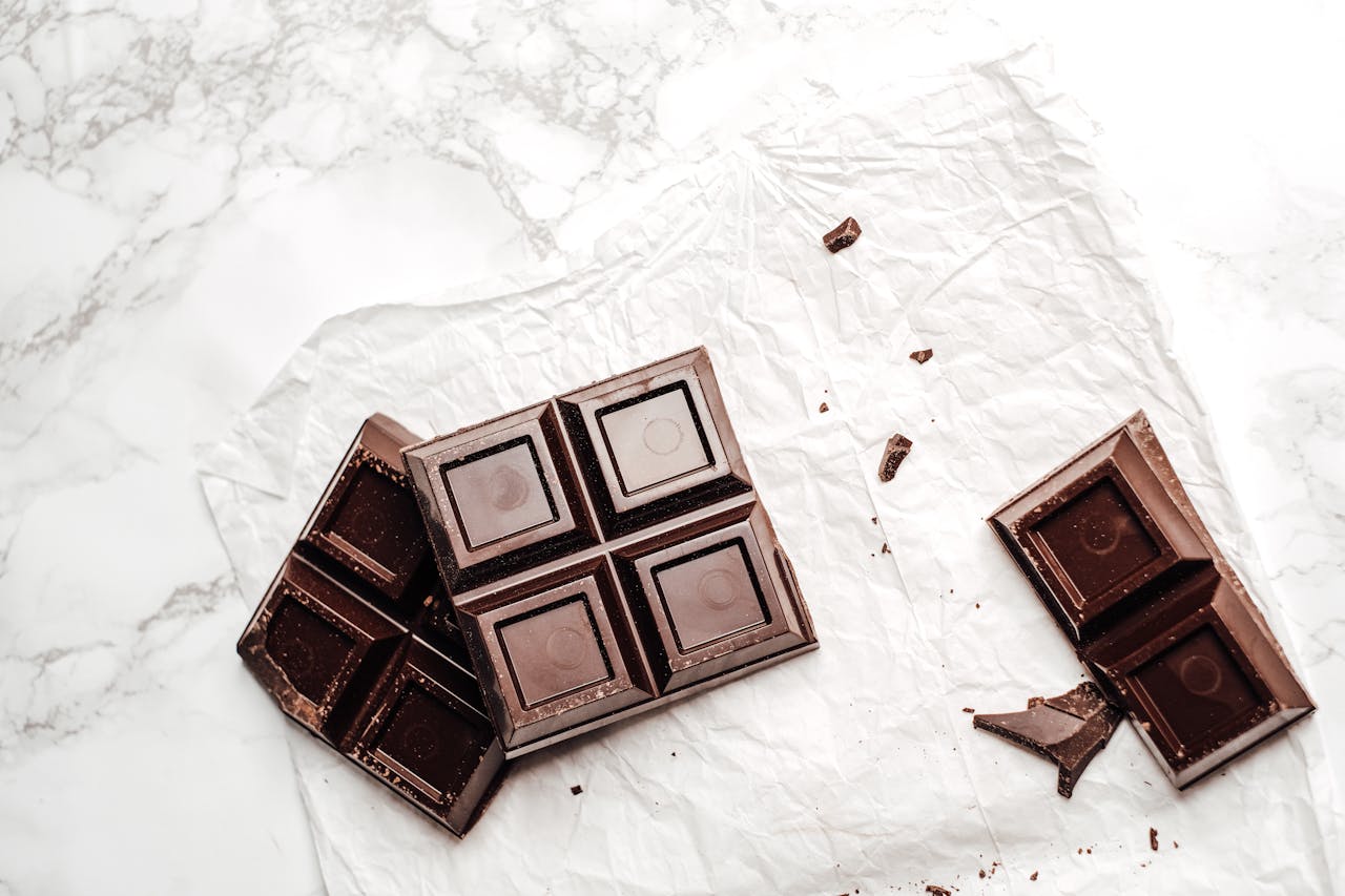 Dark chocolate bar broken into pieces on white marble surface with parchment paper and scattered chocolate crumbs